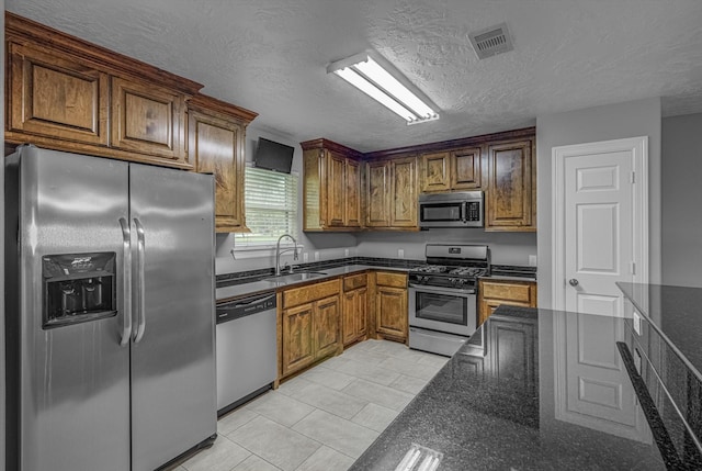2440 Olympia Dr, West Columbia, TX 77486 - kitchen with appliances with stainless steel finishes, a textured ceiling, brown cabinetry, light tile patterned flooring, and dark stone counters