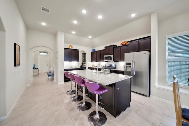 22223 Ebbets Field Dr, Spring, TX 77389 - kitchen with arched walkways, appliances with stainless steel finishes, an island with sink, light stone countertops, and dark brown cabinets