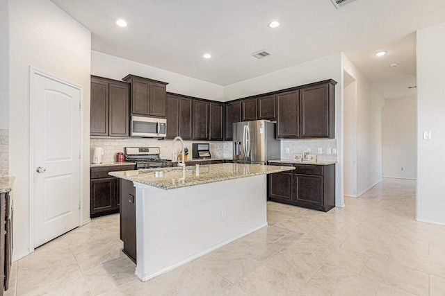 23719 Scotsman Dr, Katy, TX 77493 - kitchen featuring appliances with stainless steel finishes, backsplash, an island with sink, light stone countertops, and dark brown cabinetry