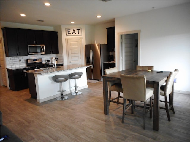 8227 Precious Passing Way, Rosharon, TX 77583 - kitchen with stainless steel appliances, an island with sink, light stone counters, dark cabinets, and light wood-style floors