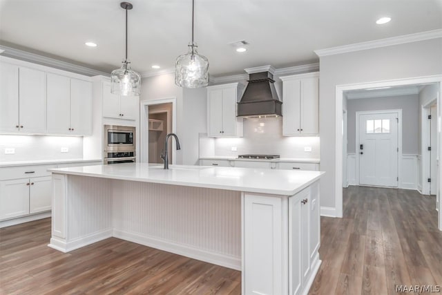 kitchen with stainless steel appliances, sink, white cabinets, custom range hood, and a center island with sink