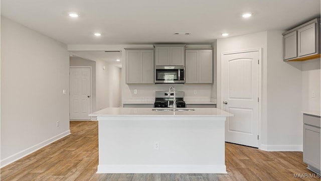 kitchen with light wood-type flooring, appliances with stainless steel finishes, a kitchen island with sink, and gray cabinetry