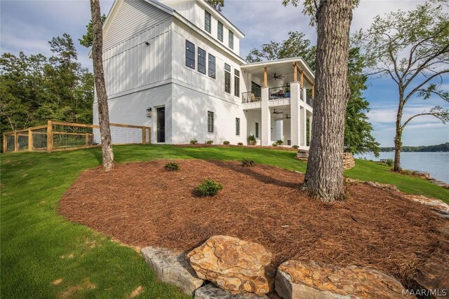 back of property with ceiling fan, a balcony, a water view, and a yard