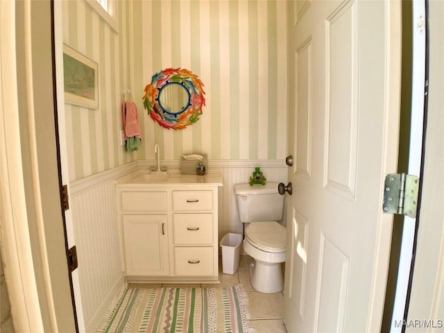 half bathroom with wallpapered walls, toilet, tile patterned flooring, and wainscoting