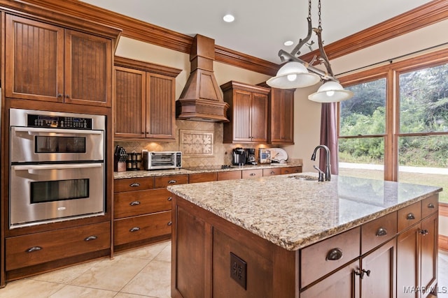 kitchen with stainless steel double oven, a sink, hanging light fixtures, custom exhaust hood, and an island with sink