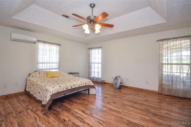 bedroom with a textured ceiling, a raised ceiling, a wall mounted AC, hardwood / wood-style flooring, and ceiling fan