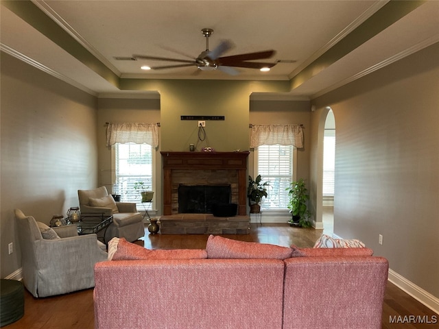 living room with ceiling fan, a stone fireplace, crown molding, and dark wood-type flooring