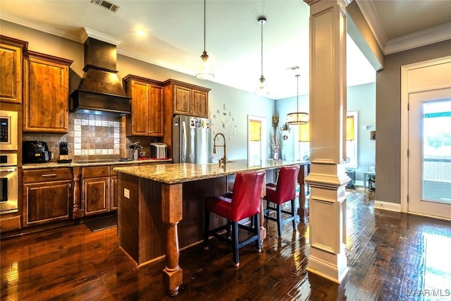 kitchen featuring a breakfast bar, hanging light fixtures, an island with sink, appliances with stainless steel finishes, and custom range hood