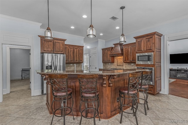 kitchen featuring premium range hood, appliances with stainless steel finishes, a kitchen island with sink, hanging light fixtures, and dark stone countertops