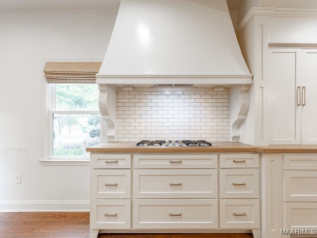 kitchen with white cabinets, stainless steel gas stovetop, custom range hood, light hardwood / wood-style floors, and decorative backsplash