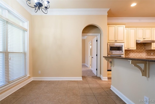 kitchen featuring stainless steel microwave, decorative backsplash, ornamental molding, cream cabinetry, and light tile patterned flooring