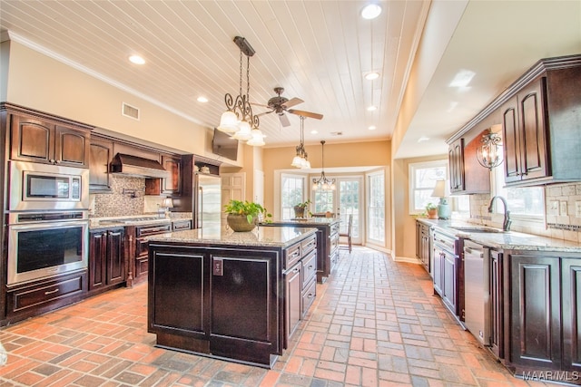 kitchen with tasteful backsplash, a kitchen island, dark brown cabinets, built in appliances, and ceiling fan with notable chandelier