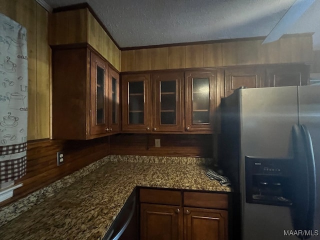 kitchen with dark stone countertops, stainless steel refrigerator with ice dispenser, wood walls, and a textured ceiling