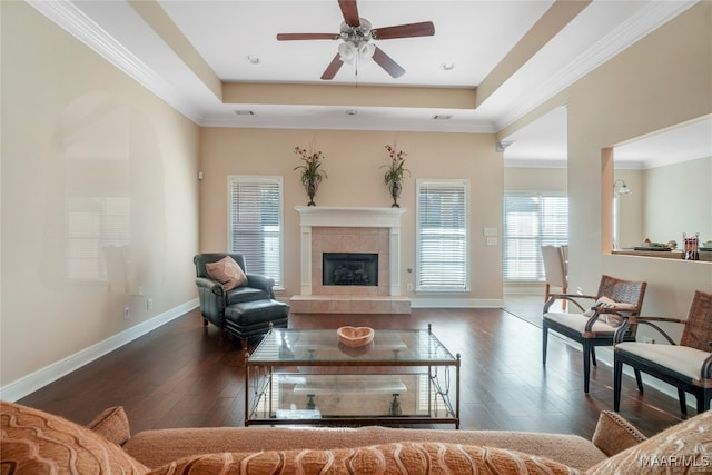 living room featuring ceiling fan, a raised ceiling, a tiled fireplace, crown molding, and dark hardwood / wood-style flooring