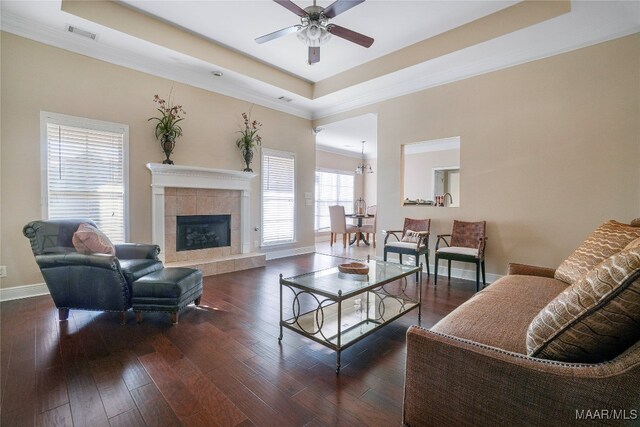 living room featuring a tray ceiling, a tiled fireplace, dark hardwood / wood-style flooring, ornamental molding, and ceiling fan