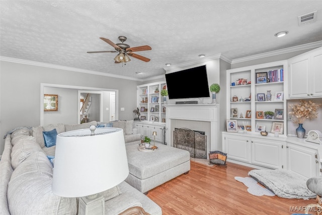 living room featuring ceiling fan, a tile fireplace, light hardwood / wood-style flooring, ornamental molding, and a textured ceiling