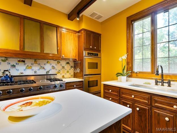 kitchen with stainless steel double oven, light countertops, a sink, beam ceiling, and visible vents