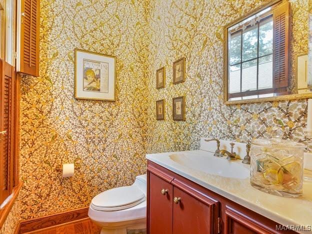 half bathroom with wood finished floors, wallpapered walls, toilet, vanity, and baseboards