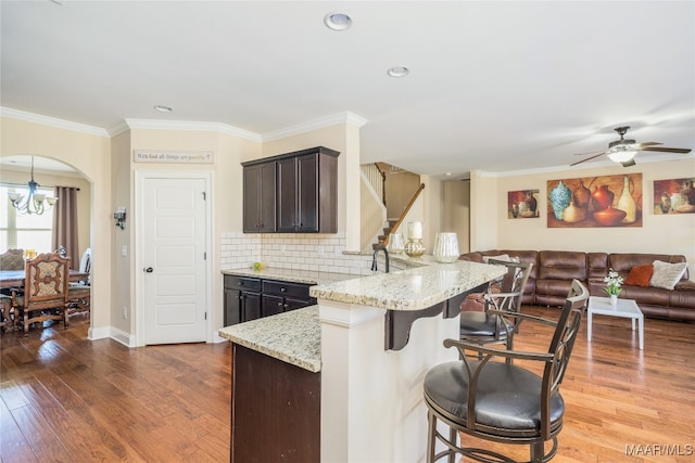 kitchen featuring hardwood / wood-style flooring, ornamental molding, light stone countertops, a kitchen bar, and ceiling fan with notable chandelier