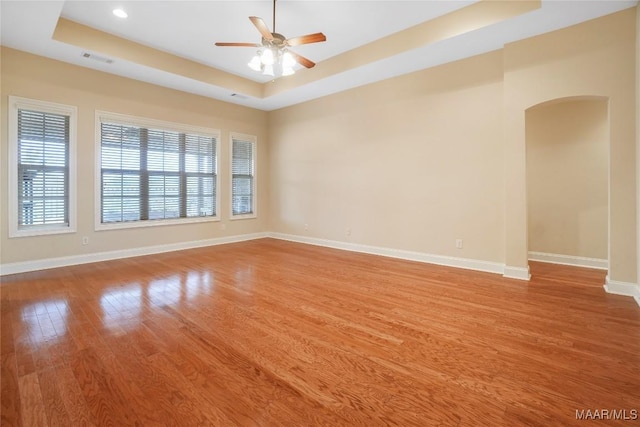 spare room with a tray ceiling, arched walkways, wood finished floors, ceiling fan, and baseboards