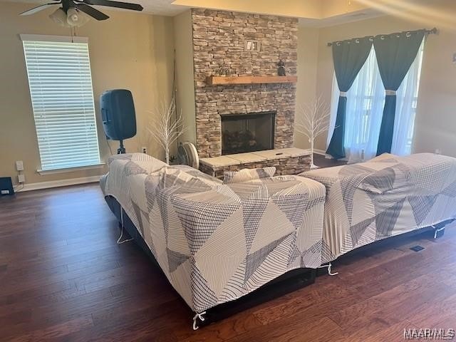 bedroom featuring a stone fireplace, multiple windows, dark wood-type flooring, and ceiling fan