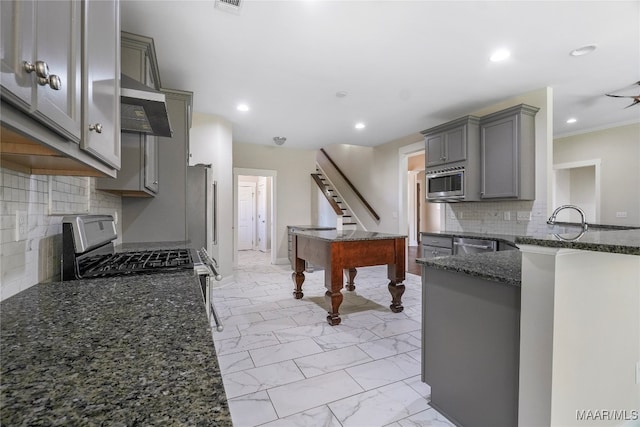 kitchen featuring range hood, backsplash, dark stone counters, gray cabinets, and appliances with stainless steel finishes