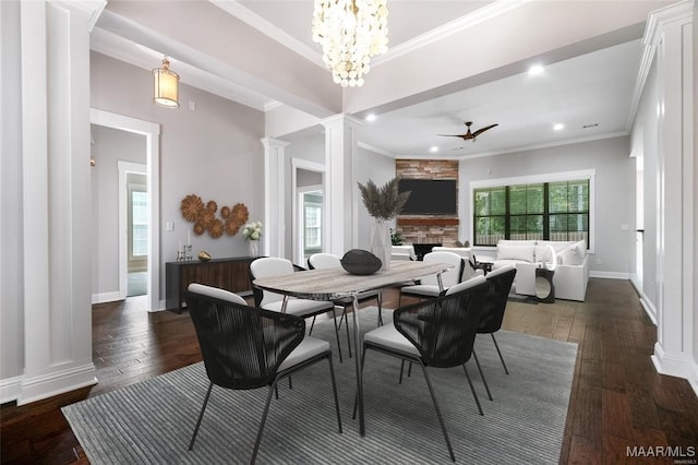 dining space featuring a stone fireplace, dark wood-type flooring, ceiling fan with notable chandelier, and ornate columns