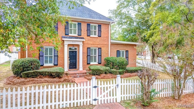 colonial inspired home featuring a fenced front yard, a gate, a shingled roof, and brick siding