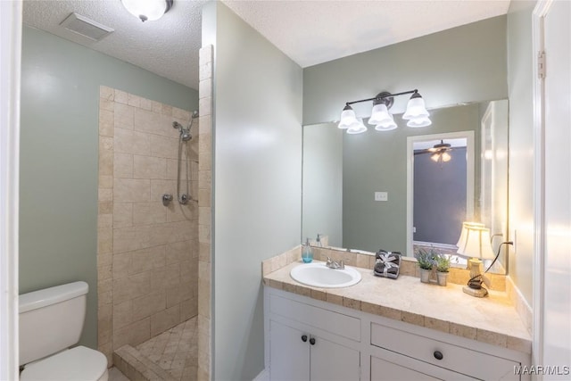 bathroom featuring vanity, ceiling fan, toilet, tiled shower, and a textured ceiling