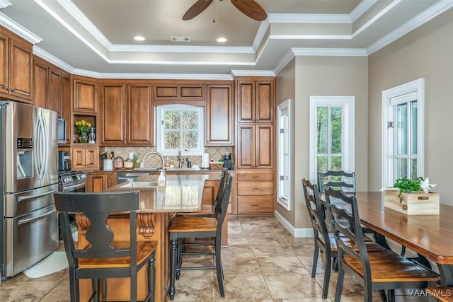 kitchen with ceiling fan, tasteful backsplash, a tray ceiling, a center island with sink, and appliances with stainless steel finishes