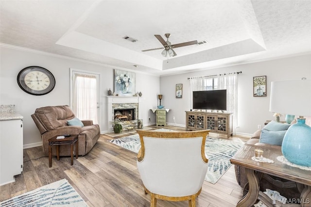 living room with a raised ceiling, a stone fireplace, ceiling fan, and light hardwood / wood-style flooring