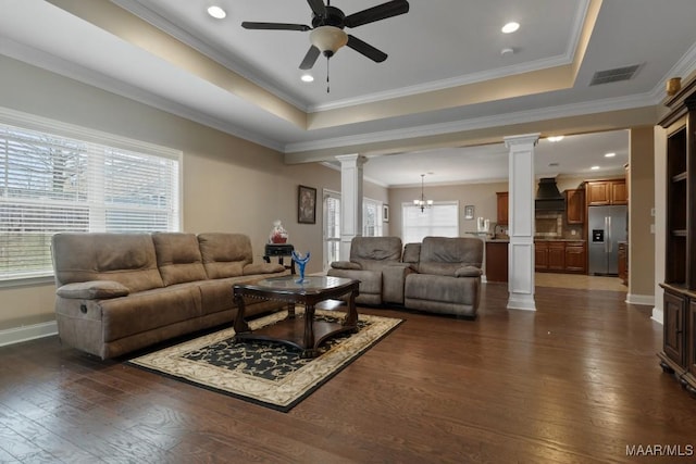 living room featuring decorative columns, ceiling fan with notable chandelier, a tray ceiling, crown molding, and dark hardwood / wood-style floors