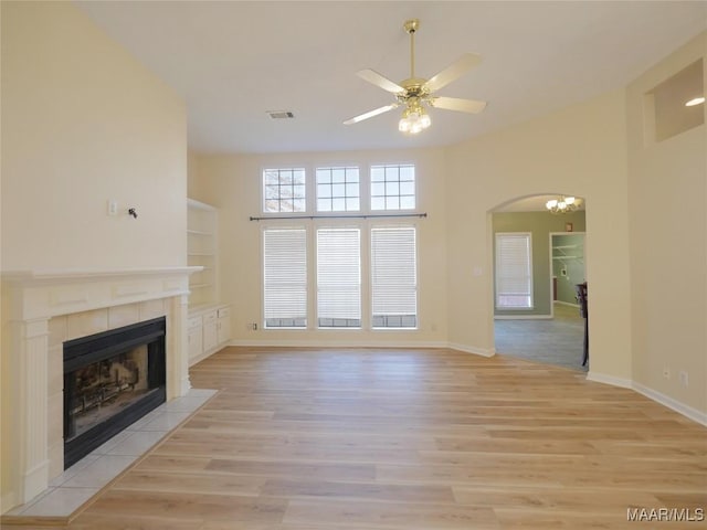 unfurnished living room featuring ceiling fan, a towering ceiling, built in features, light hardwood / wood-style floors, and a tiled fireplace