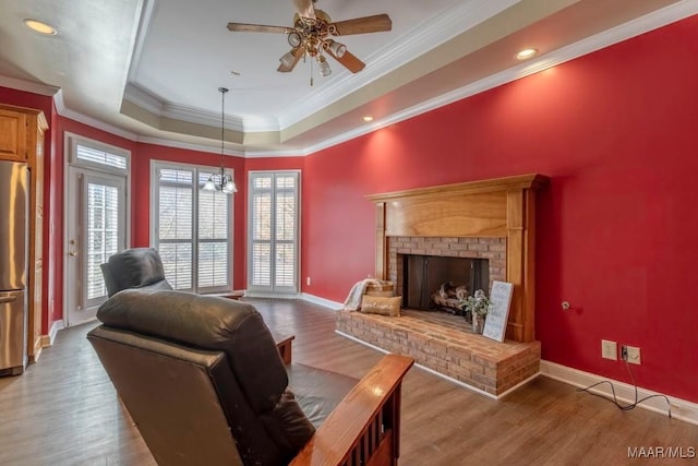 living room with a brick fireplace, a raised ceiling, wood-type flooring, ceiling fan with notable chandelier, and ornamental molding