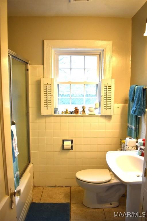 bathroom featuring tile patterned flooring, bath / shower combo with glass door, tile walls, and toilet