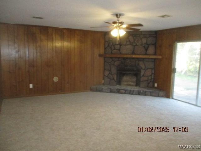 unfurnished living room with carpet floors, a stone fireplace, ceiling fan, and wood walls