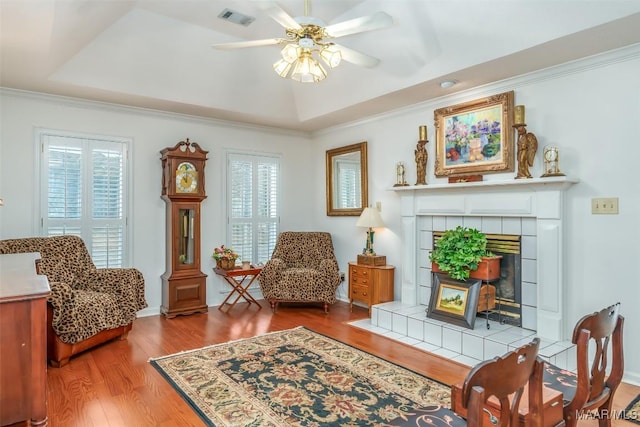 living area featuring a tray ceiling, ceiling fan, crown molding, hardwood / wood-style flooring, and a tiled fireplace