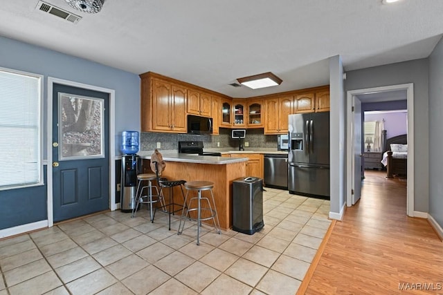 kitchen with light countertops, glass insert cabinets, brown cabinetry, a peninsula, and black appliances