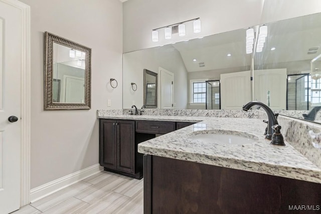full bath featuring two vanities, baseboards, vaulted ceiling, a shower stall, and a sink