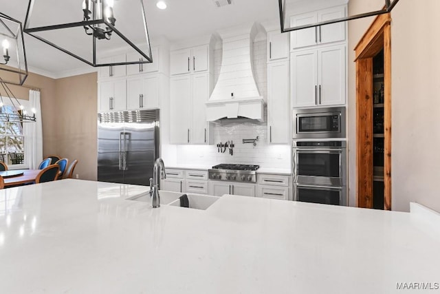 kitchen with sink, white cabinetry, built in appliances, a notable chandelier, and custom range hood