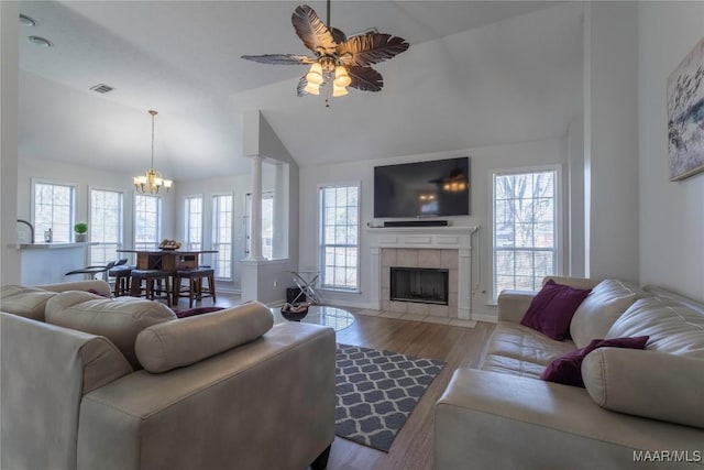 living room with decorative columns, vaulted ceiling, light wood-type flooring, a tiled fireplace, and ceiling fan with notable chandelier