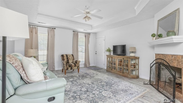 living room featuring crown molding, light hardwood / wood-style flooring, a raised ceiling, ceiling fan, and a tiled fireplace