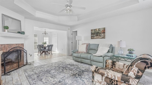 living room featuring light hardwood / wood-style floors, ornamental molding, a tiled fireplace, ceiling fan with notable chandelier, and a raised ceiling