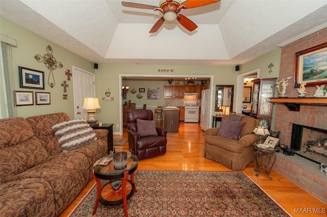 living room with ceiling fan, a tray ceiling, a brick fireplace, and light wood-type flooring