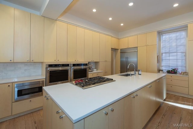 kitchen featuring sink, built in appliances, light wood-type flooring, a kitchen island with sink, and backsplash