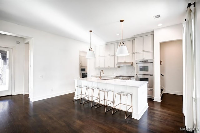 kitchen with under cabinet range hood, a sink, light countertops, an island with sink, and decorative light fixtures