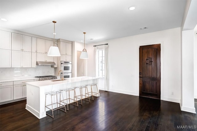 kitchen with a kitchen island with sink, stainless steel appliances, light countertops, under cabinet range hood, and pendant lighting