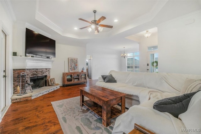 living room featuring crown molding, ceiling fan with notable chandelier, wood-type flooring, a stone fireplace, and a raised ceiling