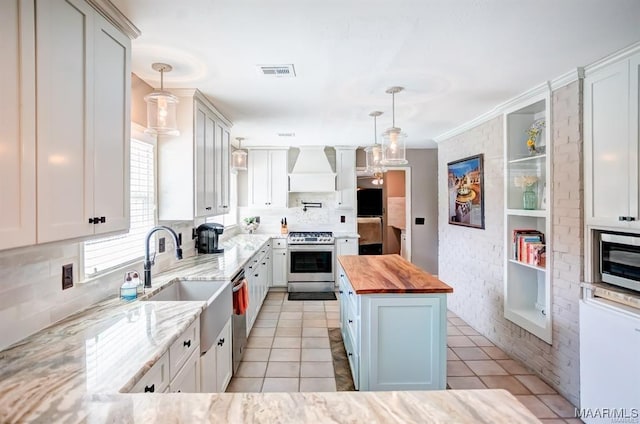 kitchen featuring premium range hood, butcher block counters, stainless steel appliances, white cabinets, and decorative light fixtures