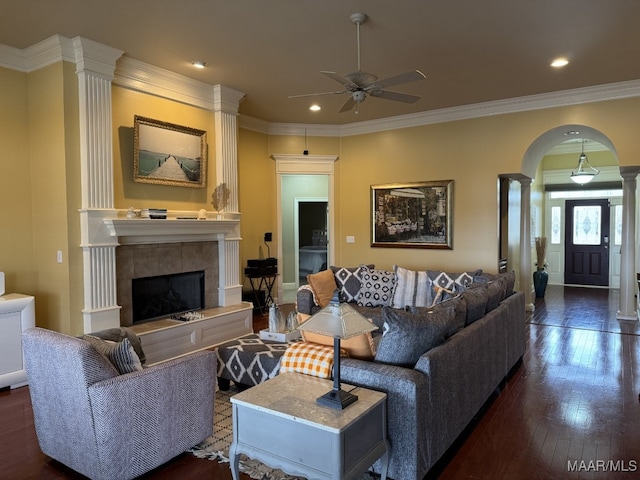 living room featuring a tile fireplace, decorative columns, ornamental molding, ceiling fan, and dark wood-type flooring